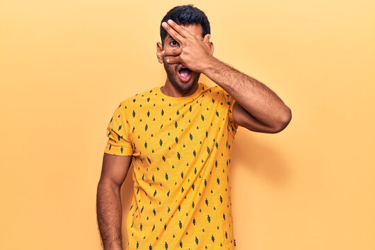 Young latin man wearing casual clothes peeking in shock covering face and eyes with hand, looking through fingers afraid