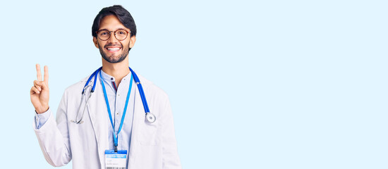 Handsome hispanic man wearing doctor uniform and stethoscope smiling with happy face winking at the...