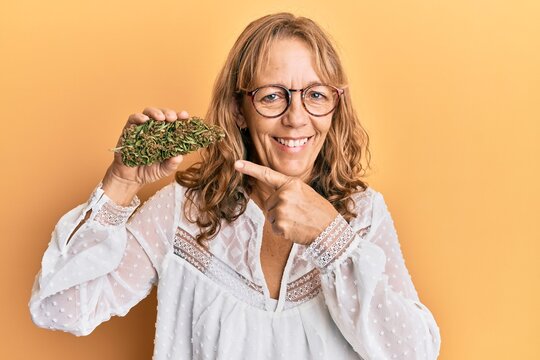 Middle age blonde woman holding medical marijuana plant bud smiling happy pointing with hand and finger
