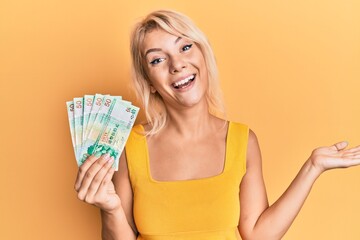 Young blonde girl holding 50 hong kong dollars banknotes celebrating achievement with happy smile and winner expression with raised hand