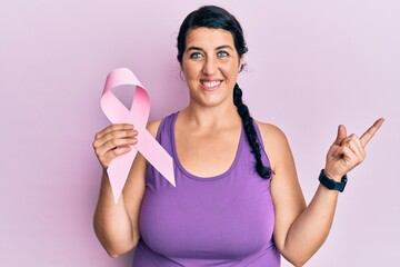 Plus size brunette woman holding pink cancer ribbon smiling happy pointing with hand and finger to the side