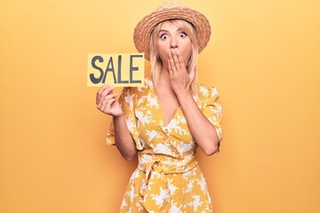 Young beautiful blonde woman on vacation wearing summer hat holding sale banner covering mouth with...