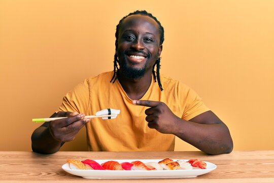 Handsome young black man eating sushi sitting on the table smiling happy pointing with hand and finger