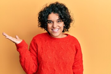 Young hispanic woman with curly hair wearing casual winter sweater smiling cheerful presenting and pointing with palm of hand looking at the camera.