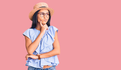 Young beautiful latin girl wearing summer hat and glasses serious face thinking about question with hand on chin, thoughtful about confusing idea