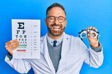 Handsome middle age man holding optometry glasses and medical exam smiling and laughing hard out loud because funny crazy joke.
