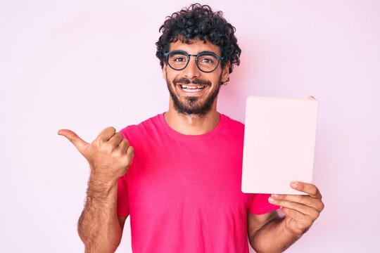 Handsome young man with curly hair and bear reading a  book pointing thumb up to the side smiling happy with open mouth