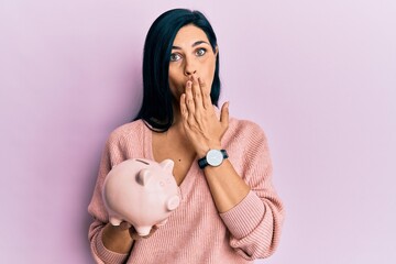 Young caucasian woman holding piggy bank covering mouth with hand, shocked and afraid for mistake....