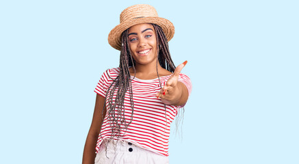 Obraz na płótnie Canvas Young african american woman with braids wearing summer hat smiling cheerful offering palm hand giving assistance and acceptance.