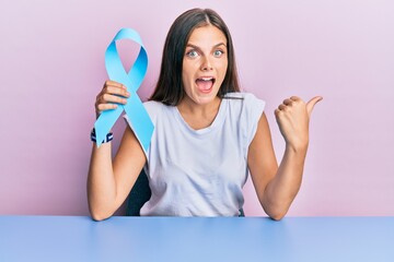 Young caucasian woman holding blue ribbon pointing thumb up to the side smiling happy with open...