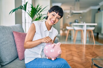Young brunette woman smiling happy putting money savings inside of piggy bank