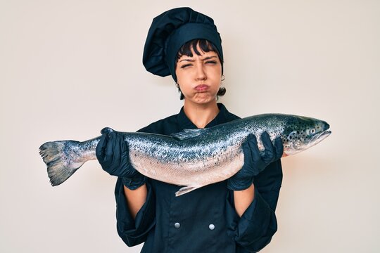 Beautiful brunettte woman professional chef holding fresh salmon fish puffing cheeks with funny face. mouth inflated with air, catching air.