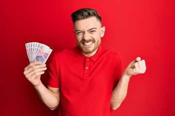 Young redhead man holding 20 polish zloty banknotes screaming proud, celebrating victory and success very excited with raised arm