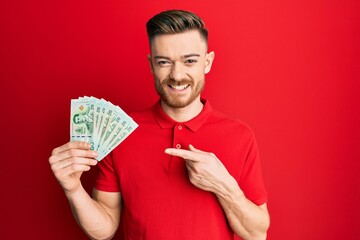 Young redhead man holding 20 thai baht banknotes smiling happy pointing with hand and finger