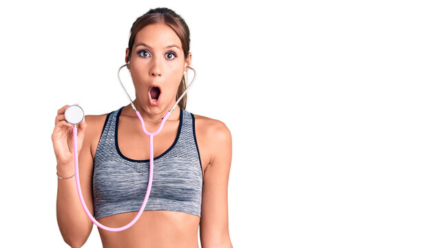 Young beautiful hispanic woman wearing sportswear holding stethoscope scared and amazed with open mouth for surprise, disbelief face