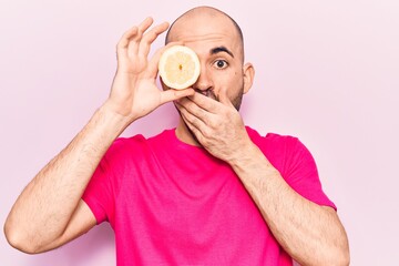 Young handsome bald man holding slice of lemon over eye covering mouth with hand, shocked and...