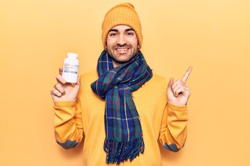 Young handsome bald man wearing winter clothes holding jar of pills smiling happy pointing with hand and finger to the side