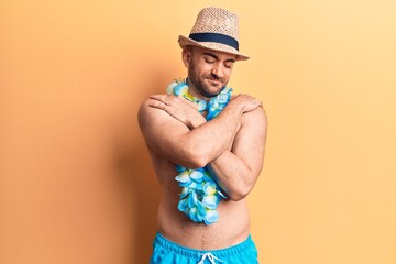 Young handsome bald man shirtless wearing swimwear and hawaiian lei hugging oneself happy and...