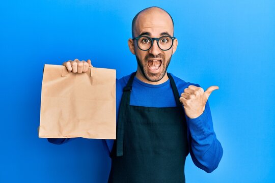 Young hispanic man wearing waiter uniform holding take away paper bag pointing thumb up to the side smiling happy with open mouth