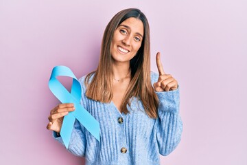 Brunette young woman holding blue ribbon smiling with an idea or question pointing finger with...