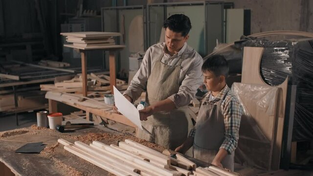 Medium shot of father and son spending time together in carpentry workshop looking at blueprint of diy furniture made of wood
