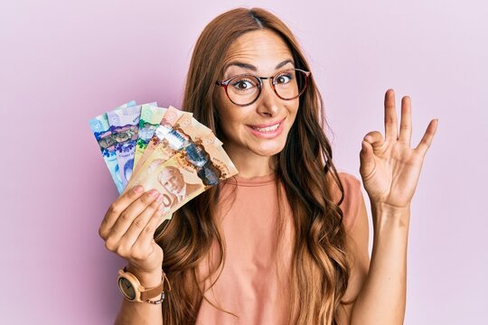 Young brunette woman holding canadian dollars doing ok sign with fingers, smiling friendly gesturing excellent symbol