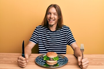 Handsome caucasian man with long hair eating a tasty classic burger winking looking at the camera...