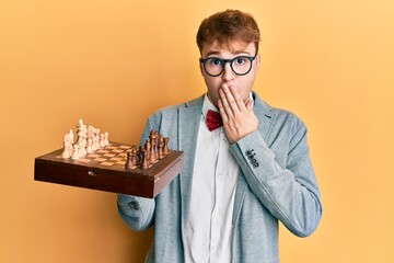 Young caucasian man wearing glasses holding chess board covering mouth with hand, shocked and...