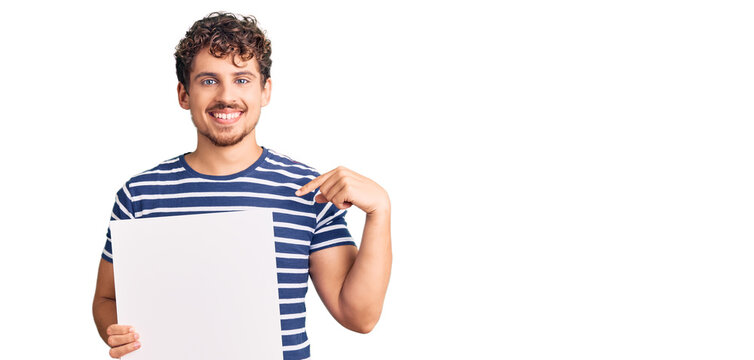 Young handsome man with curly hair holding blank empty banner pointing finger to one self smiling happy and proud