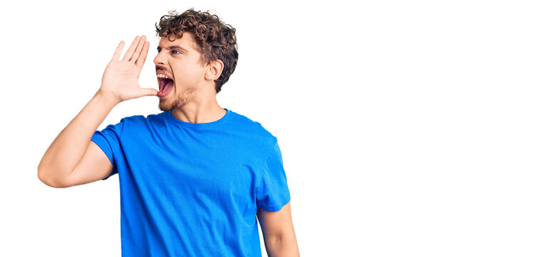 Young handsome man with curly hair wearing casual clothes shouting and screaming loud to side with hand on mouth. communication concept.