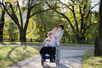 マスクをつけて車椅子で公園を散歩する日本人シニア男性と介助する妻