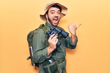 Young hispanic man wearing explorer clothes holding binoculars pointing thumb up to the side smiling happy with open mouth