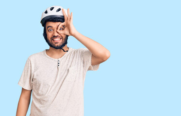 Young hispanic man wearing bike helmet smiling happy doing ok sign with hand on eye looking through fingers