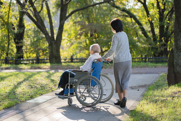 マスクをつけて車椅子で公園を散歩する日本人シニア男性と介助する妻