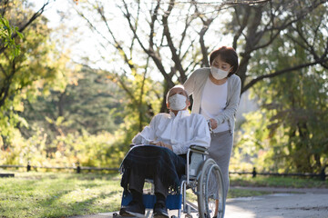 マスクをつけて車椅子で公園を散歩する日本人シニア男性と介助する妻