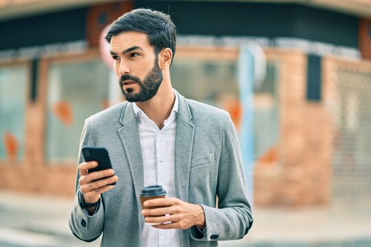 Young hispanic businessman with serious expression using smartphone and drinking coffee at the city.