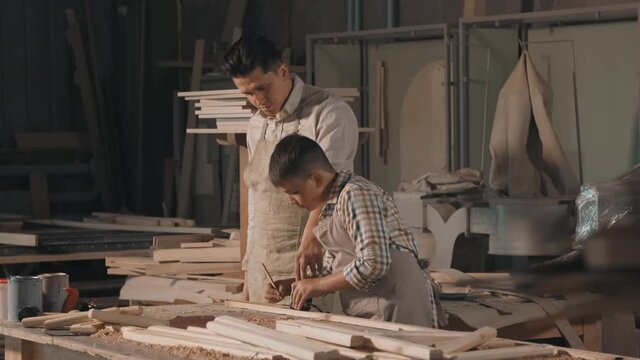 Medium shot of caucasian man and his son working in carpentry workshop cutting wood with jigsaw for making diy furniture