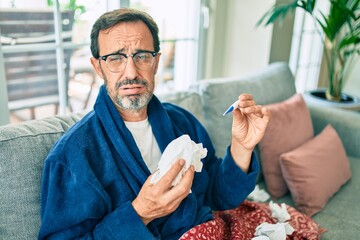 Middle age man feeling sick with cold and fever at home, ill with flu disease sitting on the sofa