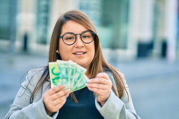 Young hispanic plus size businesswoman smiling happy holding israel shekels banknotes at the city.