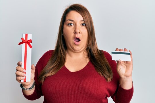 Beautiful brunette plus size woman holding gift and credit card afraid and shocked with surprise and amazed expression, fear and excited face.