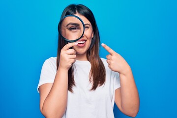 Young beautiful brunette woman using magnifying glass over isolated blue background smiling happy pointing with hand and finger