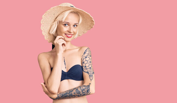 Young blonde woman with tattoo wearing bikini and summer hat looking confident at the camera with smile with crossed arms and hand raised on chin. thinking positive.