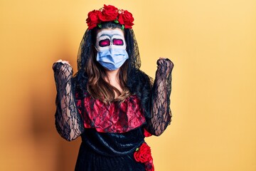 Young woman wearing day of the dead costume wearing medical mask very happy and excited doing winner gesture with arms raised, smiling and screaming for success. celebration concept.