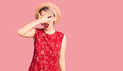 Young beautiful blonde woman wearing summer hat peeking in shock covering face and eyes with hand, looking through fingers with embarrassed expression.