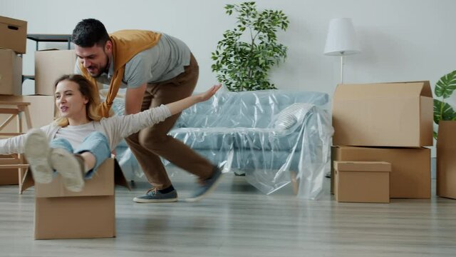 Slow motion of happy family husband and wife enjoying funny activity riding boxes during relocation to new modern flat. Happiness and emotions concept.