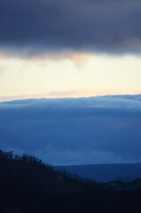 Storm clouds are gathering on the horizon at dusk above a forest filled valley. Clear sky is above the storm, with dark clouds tinged with the colours of sunset.