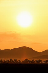 Picture of a sun setting behind a dense forest area followed by mountains.	