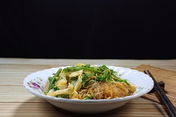 Fried and stirred instant noodle mixed with vermicelli and local herbal vegetable on the plate. Fusion vegetarian menu in Asia restaurant. 