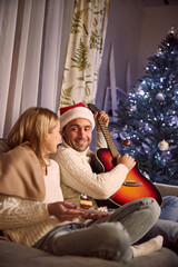 Sweet smiling young couple eating cake and listening play the guitar in living room in Christmas Eve