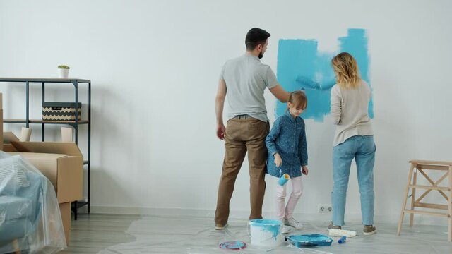 Mother father and little daughter are painting apartment wall decorating room together using paddle roller and bright paint. People and renovation concept.